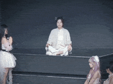 a woman in a white dress sits on a ledge with her legs crossed while another woman sings into a microphone