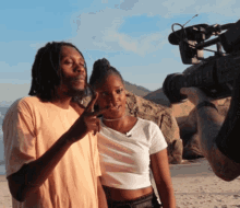 a man and a woman are standing on a beach while a cameraman takes a picture
