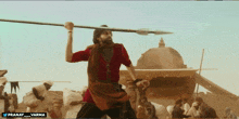 a man in a red shirt is holding a large spear in front of a group of people and a sign that says pranay
