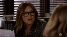 a woman wearing glasses and a polka dot shirt is talking to another woman with the nbc logo behind her