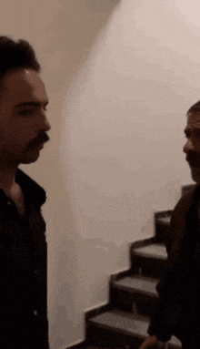 two men with mustaches are standing next to each other in front of a staircase .