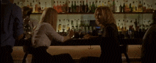 two women are sitting at a bar talking to each other with bottles of alcohol behind them