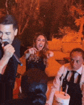 a man is singing into a microphone while a woman laughs and a waiter stands behind her .