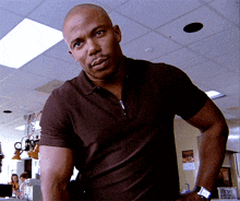 a bald man wearing a brown polo shirt and a watch stands in a room