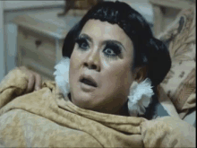 a drag queen is sitting on a bed with a blanket on her head .