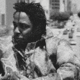 a black and white photo of a man with dreadlocks wearing a camouflage jacket