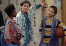three boys are standing in a locker room talking to each other while one of them is holding a basketball .