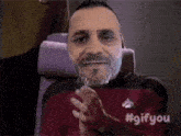 a man with a beard is sitting in a chair with his hands folded in front of his face .