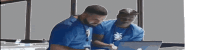 two men in blue shirts look at a laptop