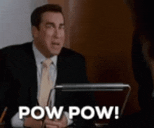 a man in a suit and tie is sitting at a desk with a laptop computer and says pow pow .