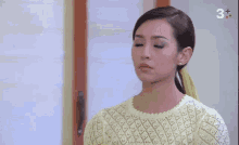 a woman in a yellow sweater stands in front of a window with a 3+ sign on it