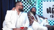 two men in white suits are sitting next to each other in front of a sign that says he