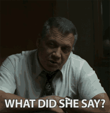 a man in a white shirt and tie is sitting at a table and says " what did she say "