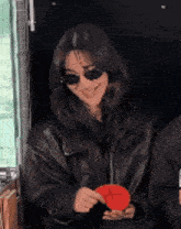 a woman wearing sunglasses and a leather jacket is holding a red object