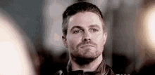 a man with a beard is wearing a leather jacket and looking at the camera .