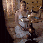 a woman in a white dress sits on the floor next to a table with a book on it