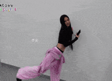 a woman in pink pants and a black top is kicking her leg up in front of a wall that says meovv myself