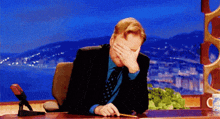 a man in a suit and tie is sitting at a desk covering his face with his hand .
