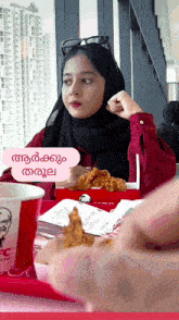 a woman in a hijab is sitting at a table with a box of fried chicken and a kfc cup