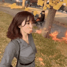 a woman is blowing leaves in the air in a park while a man sits on a bench behind her .
