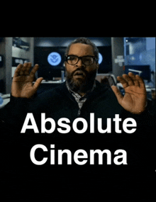 a man with glasses and a beard stands in front of a poster that says absolute cinema