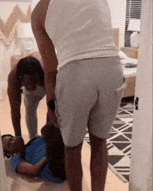 a man in a white tank top and grey shorts is standing next to another man laying on the floor