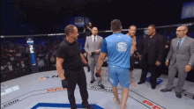 a man in a blue shirt stands in a boxing ring surrounded by a group of men