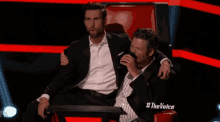 two men are sitting next to each other on a red chair with #thevoice written on it