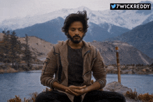 a man sits in a lotus position with his eyes closed in front of mountains and a lake