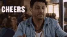 a man in a blue shirt is sitting at a table in a restaurant with the words `` cheers '' written on the screen .