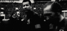 a black and white photo of two men standing next to each other in front of a crowd .