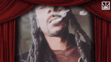 a man with long dreadlocks is smoking a cigarette behind a red curtain .