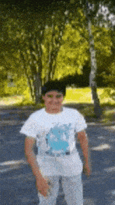 a young boy wearing a white shirt and white pants is standing in front of trees in a park .