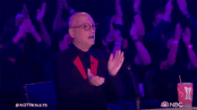 a man wearing glasses is clapping his hands in front of a crowd of people .