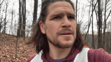 a man with long hair and a beard is wearing a white vest in the woods