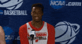 a man wearing a consin basketball jersey stands in front of a ncaa logo