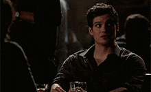 a man is sitting at a table in a dark room with a glass of water in his hand .