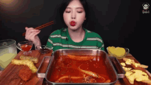 a woman in a green striped shirt is eating a tray of food with chopsticks