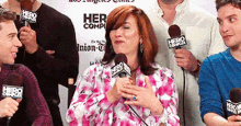 a woman in a pink shirt is speaking into a microphone in front of a sign that says her comp