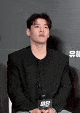 a man in a black jacket is sitting in front of a microphone with a sign that says ' ae ' on it