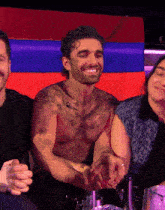 a shirtless man with a tattoo on his chest smiles while sitting at a table
