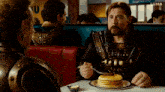 a man in armor is eating pancakes in a diner while another man watches .