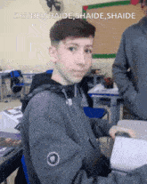 a boy is sitting at a desk in a classroom with the words shaide shaide shaide shaide written above him