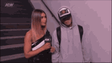 a woman is standing next to a man wearing a hoodie and a hat .