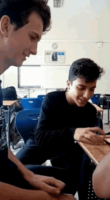 two young men are sitting at desks in a classroom with a clock on the wall
