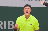 a man wearing a yellow adidas shirt holds a tennis racquet in front of a sign that says aries