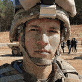 a soldier wearing a helmet looks at the camera with a group of people walking in the background