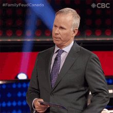 a man in a suit and tie is holding a piece of paper in front of a sign that says family feud canada