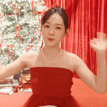a woman in a red dress with a pearl necklace is waving in front of a christmas tree