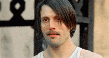 a man with long hair and a beard wearing a white tank top is looking at the camera .